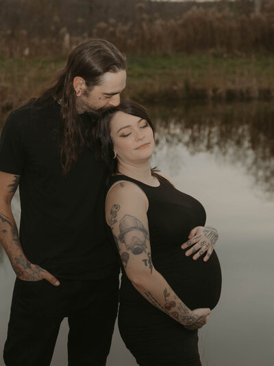 A photograph of a couple. the woman is pregnant and leaning into her partner with her eyes closed, while she embraces her growing belly. the backdrop of their pond against the golden hour sky in western new york. Photo by Dash and Boheme Photography. 