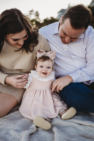 A smiling baby girl in a pink dress with a matching bow sits between her proud parents during a milestone family photography session in Connecticut. Captured by Nicole Mele Photography, celebrating love, joy, and baby’s precious early moments.