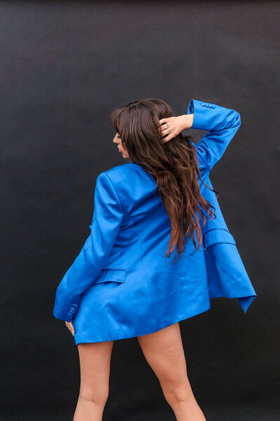 Confident woman with long hair posing in an oversized bright blue blazer against a black studio backdrop for creative business lifestyle photos.