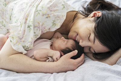 A new mother wearing a flower print dress brings her new baby close to her as they cuddle together on the bed during a newborn photo shoot with Jess Morgan Photography