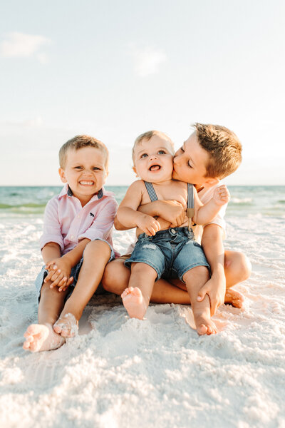 Family Photographer, a young family of four sit together at the beach, mom holds baby