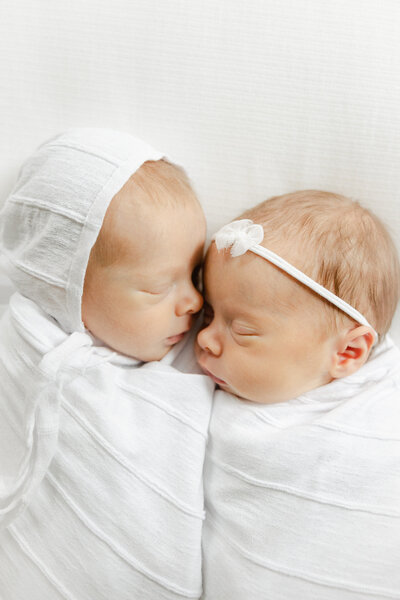 twin newborns swaddled in white lie sleeping next to eachother with their faces turned to eachother, carmel newborn photographer