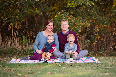 A family smiles sitting on a picnic blanket in a green grassy field.