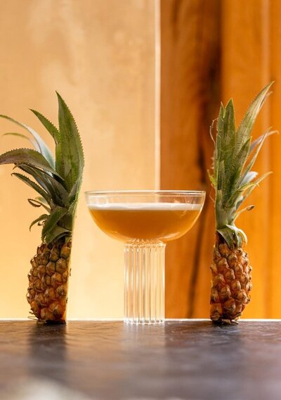 Elegant coupe glass with a golden rum cocktail flanked by two halved pineapples standing upright on a dark wooden surface.