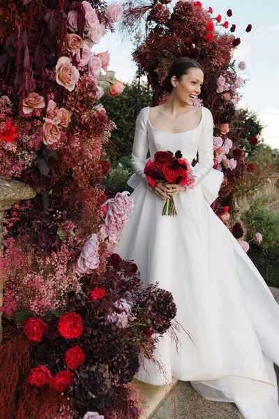 Bride in a Reem Acra wedding gown smiles beside a lavish floral arrangement in rich hues of red and pink at her Euridge Manor wedding, planned by ByChenai.