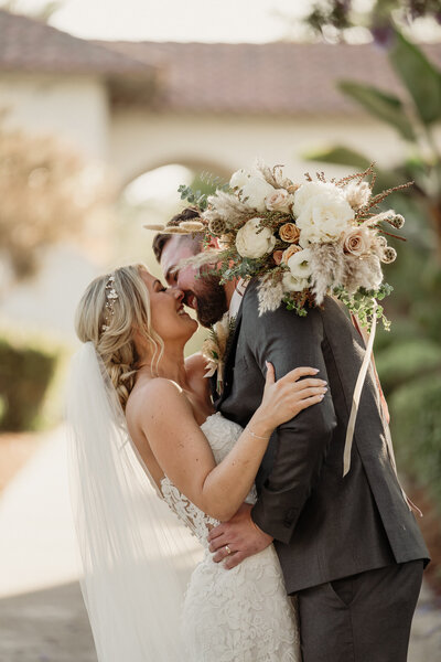 groom leans in to kiss his bride . bride is in a white lace dress with a long veil holding a brown and dusty rose boho wedding bouquet with long ribbons