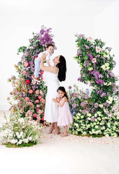 A woman in a white sleeveless dress joyfully lifts a young boy in light blue pants, while a little girl in a pink dress smiles below, all framed by colorful floral columns.