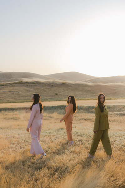 Editorial branding portrait of three women in pastel and earth-toned suits walking through a golden field at sunset, styled for a modern, fashion-forward brand shoot.