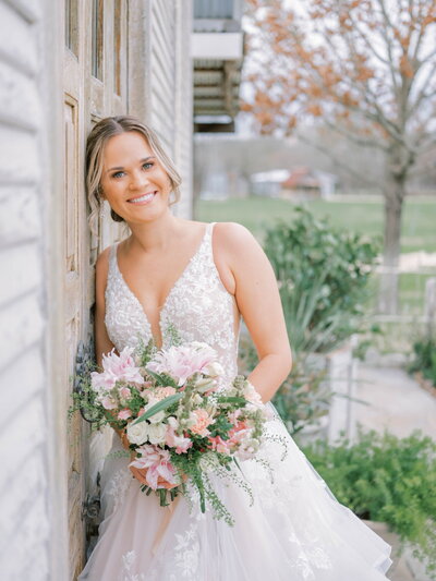 Bride in floral lace gown smiles, holding pink and green bouquet by rustic wooden wall. Her natural glam wedding makeup enhances the garden scenery backdrop, a testament to editorial bridal makeup artistry.