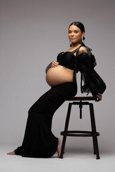 A confident pregnant woman poses in a chic black maternity outfit, sitting on a modern stool against a minimalist backdrop. Captured in a luxury Dallas maternity photography session, highlighting strength and beauty.