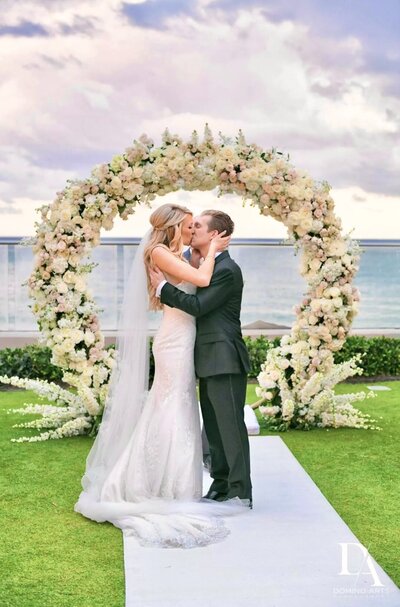 Bride and Groom kissing under thick round floral arch in front of ocean