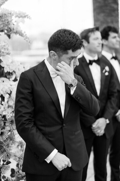 Groom wipes away tears while standing with groomsmen during an outdoor ceremony, captured in black and white