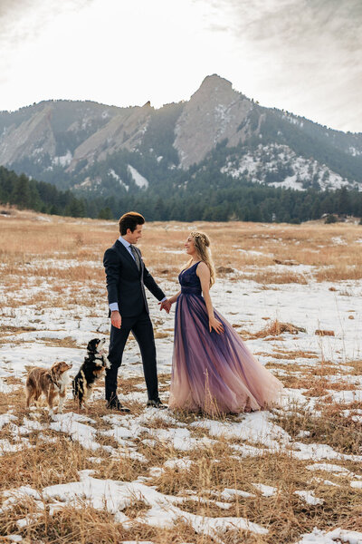 The couple is standing in snowy grass, holding hands and looking at each other. There is a mountain behind them with green trees. 