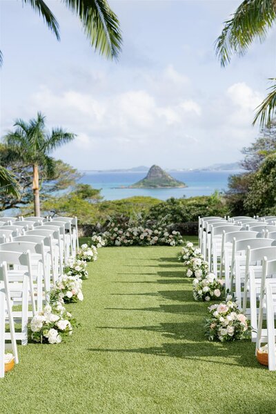 An outdoor wedding ceremony venue with white chairs and flowers lining the aisle