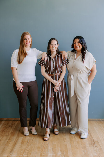 Bridget Mary Wedding and Events Team stand on a wooden floor with a grayish blue wall behind them. Alicia wears brown pants and a white shirt with her arm perched on Bridget's shoulder. Bridget stands in the middle wearing a brown and white jumpsuit and her hand holding her opposite wrist in front of her. Vanessa stands in a cream-colored jumpsuit with her hand on Bridget's shoulder.