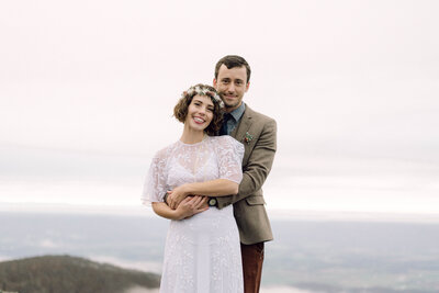 Cinematic wide shot of the couple embracing on the summit, with soft focus and natural light.
