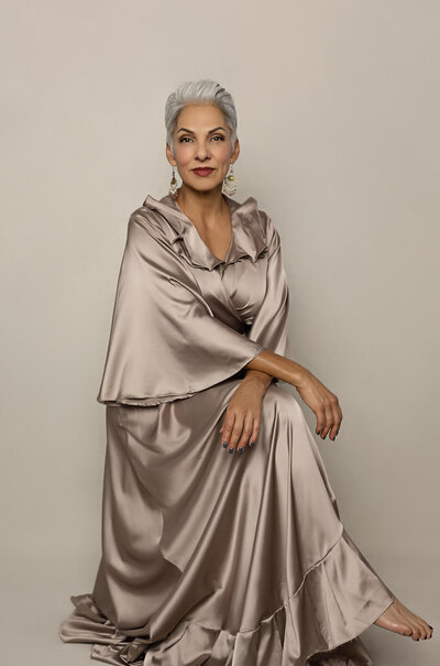 Mature woman is seated in an elegant pose and wearing a satin long dress