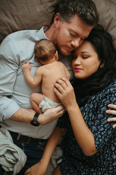 parents are cuddling their newborn at home , natural light spilling in from a window. 