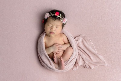 Peaceful sleeping newborn baby girl wrapped in soft pink fabric wearing pink and red floral crown photographed on matching pink textured backdrop
