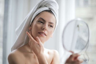 woman with hair wrapped in a towel inspecting her facial skin in a handheld mirror