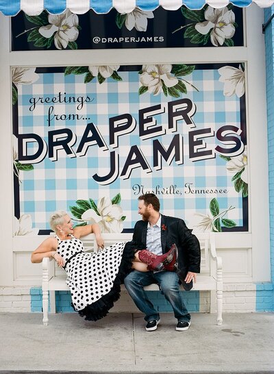 1950s pinup bride wearing a white polka dot dress and red cowboy boots and rockabilly groom wearing jeans and a black silk suit jacket holding a bright red bouquet relaxing in front of Draper James in Nashville