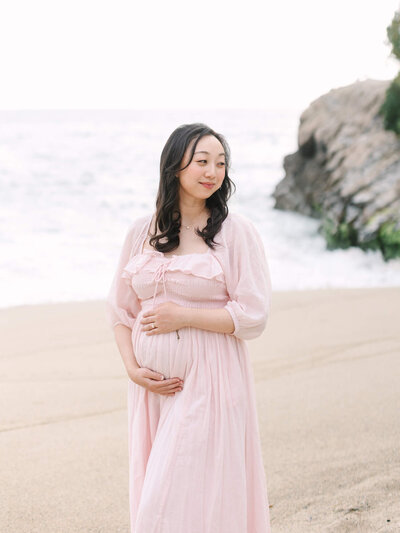 maternity photo of pregnant woman wearing a pink dress holding her belly at the beach