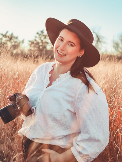 very warm sunset photo of Massachusetts Wedding Photographer Avid Aperture Photography wearing a white long-sleeve blouse and black wide-rim hat holding her camera in a tall grassy field located at Sapowet Marsh in Tiverton, Rhode Island