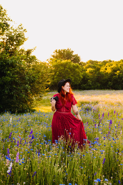Woman looking to right side in long red dress and black hat in garden with blue and purple flowers and trees. In her hand she holds a camera and holds a part of the dress