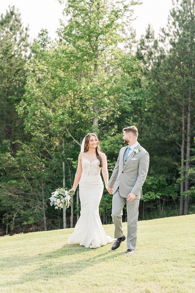 A couple holding hands while walking on their wedding day in front of a row of trees, as the bride holds her bouquet of flowers. 