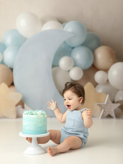 one year old boy in blue onesie grabbing and eating cake for first birthday cake smash photoshoot