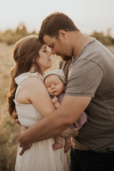 A couple is resting their foreheads on each other, while their newborn girl is nestled in between them.