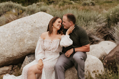 A couple sitting on a boulder while the wife holds her husband's face and smiles at him.
