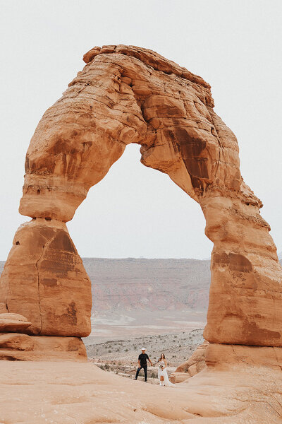 Bride and groom in cowboy and cowgirl hats walking through the sandy trail leading to Sand Dune Arch in Arches National Park