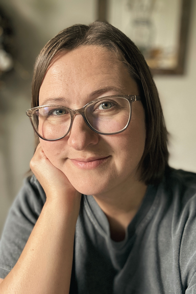 Kasey sits resting her face in her hands with a soft smile, wearing glasses and a gray shirt.