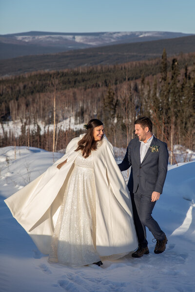 A bride wearing a white dress and a white cape twirls next to her groom as they stand in the snow on a winter day in Fairbanks Alaska.