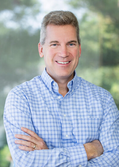 Man in a blue checkered shirt with grey hair smiling against a green background