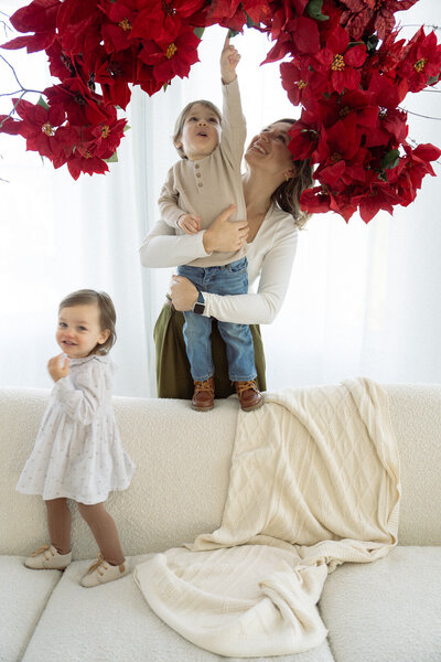 In studio photography session with mom holding son and little toddler girl walking across a modern cream colored sofa