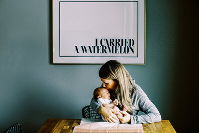 mom kisses newborn baby on forehead while sitting at dining room table