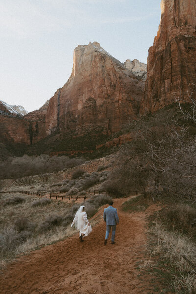 Couple in their wedding attire walking on a red dirt trail in Zion. The mountains are in the back ground while they are small in the foreground. 