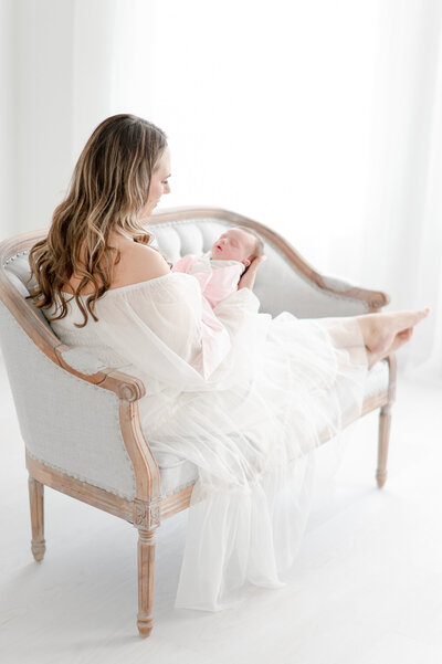 Mother in white dress sits on a gray settee in Nashville newborn photography studio