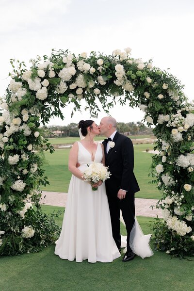 Bride and groom sharing a kiss under round floral arch with white flowers