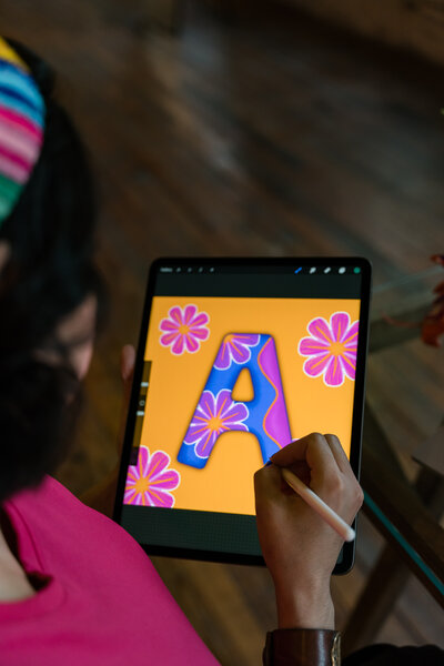 The world of digital art as the founder of Soleil Vida Studio designs a captivating piece on her iPad. Against an orange backdrop, witness the creation of the letter A adorned with pink flowers, skillfully crafted with a black digital pencil.