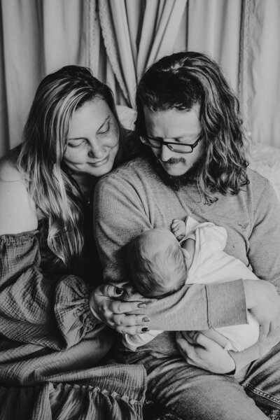 Black and white photo of mom and dad sitting on bed looking down at baby boy in lifestyle studio.