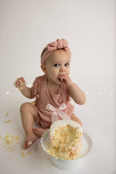 Baby girl in a pink outfit with a bow headband, enjoying a cake smash with crumbs scattered around.