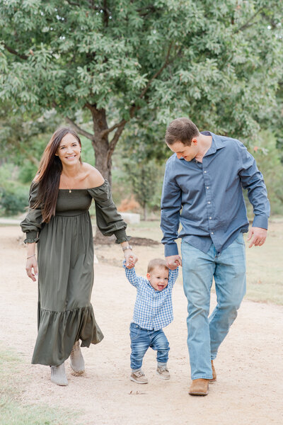 A family of three walks down a sandy path at Katherine Fleischer Park. The mother and father hold their little boy's hand who stands in between them. The little boy is laughing in this candid shot captured by a photographer specializing in family portraits in Austin, TX