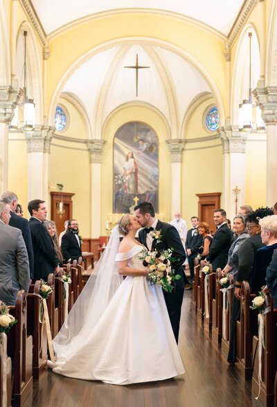 A couple dressed in wedding attire shares a kiss at the altar of a beautifully decorated church. Guests stand and watch from the pews, under an arched ceiling with stained glass windows and elegant lighting.