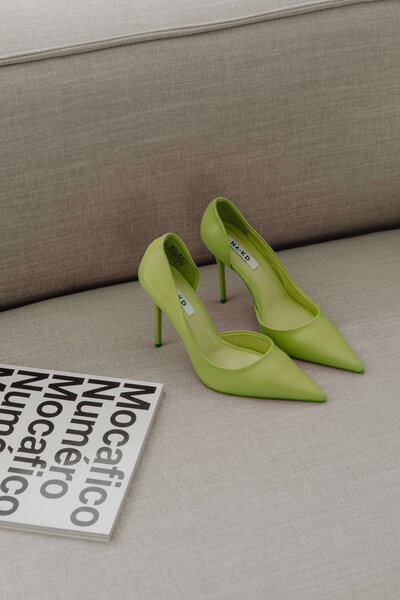 Green high-heeled shoes are placed on a beige sofa next to a book titled "Mocãfico Numero," an intriguing read.