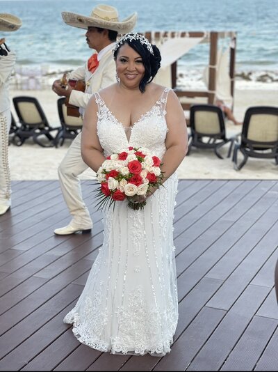 A joyful bride in an elegant lace wedding dress holding a red and white bouquet, with a mariachi musician playing in the background at a beachside wedding in Mexico.