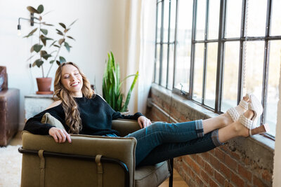 Woman sitting in a Bay Area loft sitting in a olive green chair with her legs up on the window ledge and smiling.