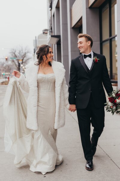 A happy bride and groom walking hand in hand along a city street in Illinois, the bride wearing a long white dress and a fur coat, the groom in a black tuxedo.